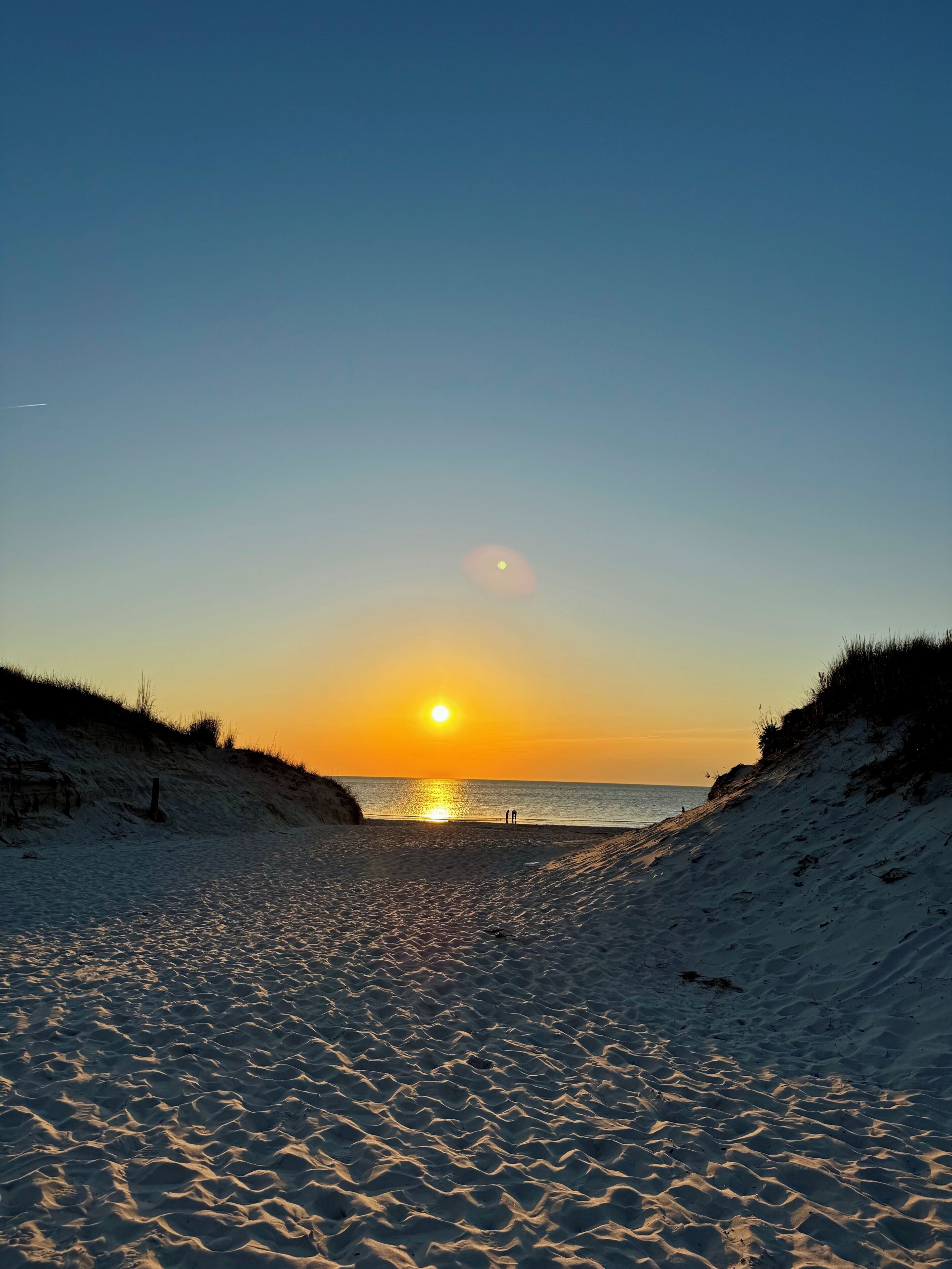 A beach with a sunset, sandy dunes, and a clear sky.