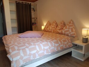 A bed with a geometric patterned bedspread, a single pink pillow, matching orange pillows, a bedside lamp, and a bookshelf with books. - Schneevogul, (Grächen), 1114 by Interhome (Grächen)