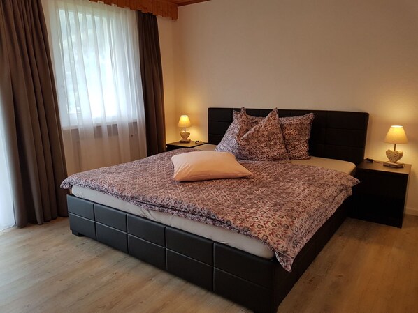 A bedroom with a large bed, two bedside tables with lamps, a window with curtains, and a wooden floor. - Schneevogul, (Grächen), 1114 by Interhome (Grächen)