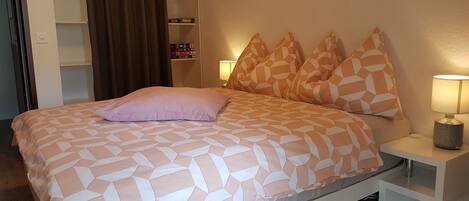 Une chambre à coucher avec un lit, une table de chevet, une lampe, une bibliothèque et un rideau.