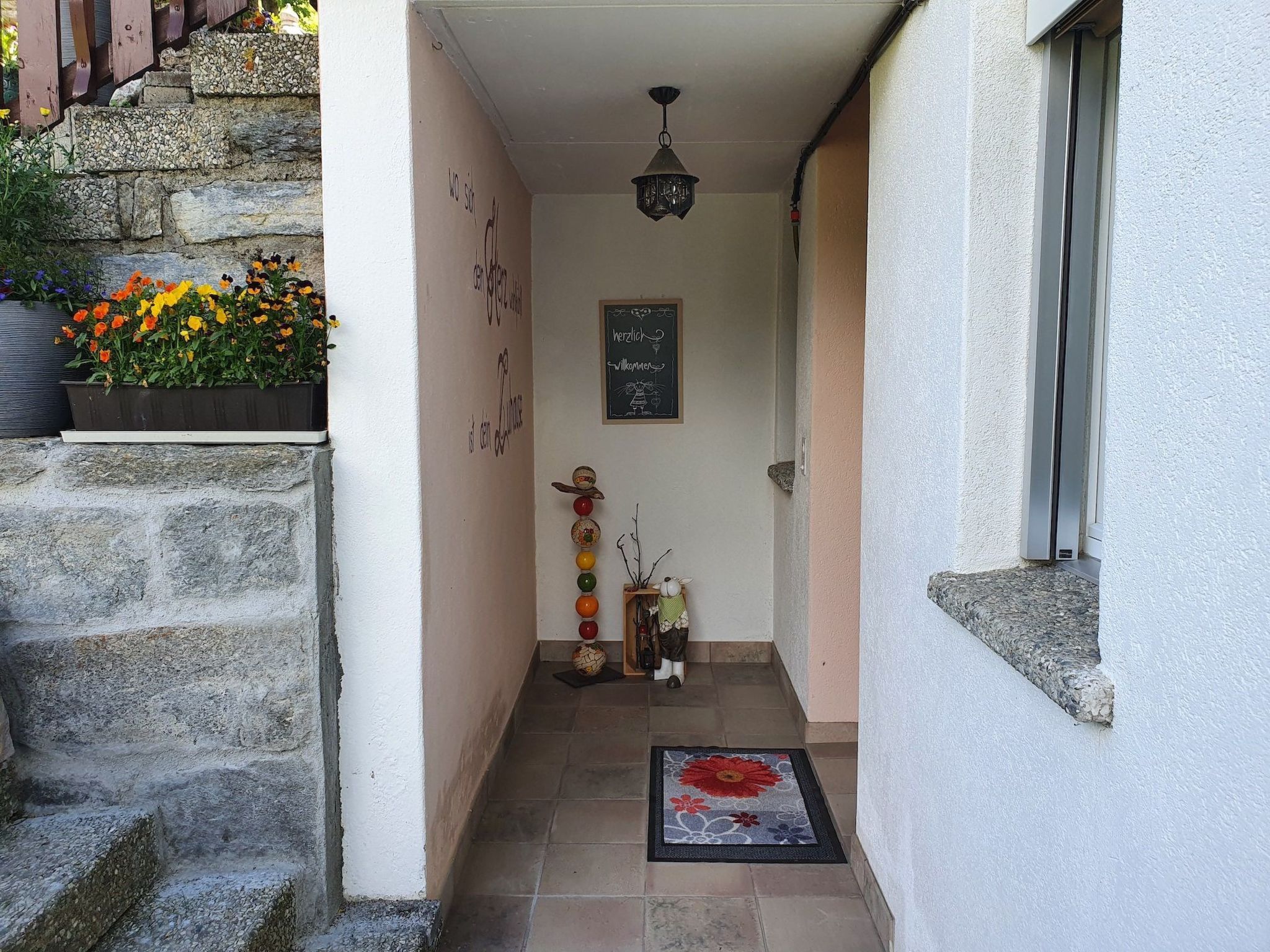 Un couloir étroit, avec un mur de pierre d’un côté et un mur blanc de l’autre, qui mène à une porte ornée d’une pancarte de bienvenue.