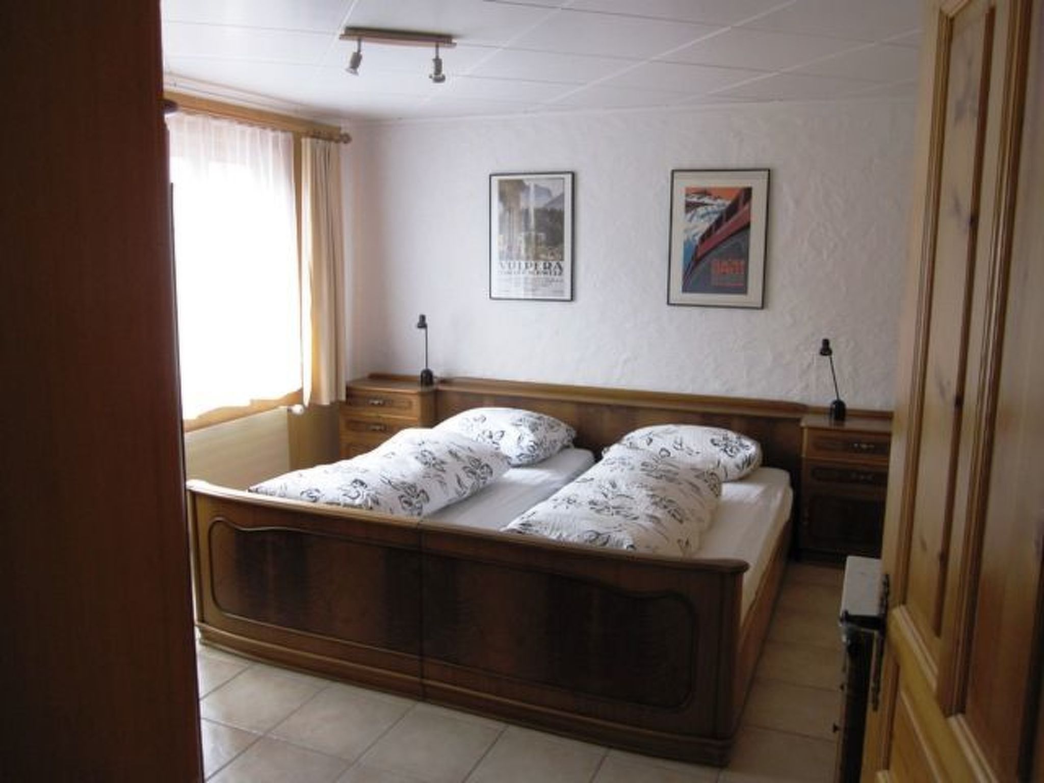 Ein Schlafzimmer mit einem hölzernen Bett, zwei Nachttischen, einem Fenster mit Vorhängen und zwei gerahmten Bildern an der Wand.
