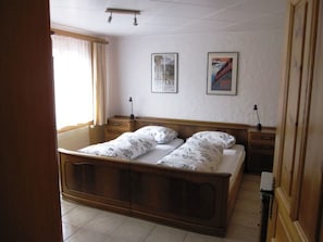 A bedroom with a wooden bed, two bedside tables, a nightstand, a window with curtains, and two framed pictures on the wall. - Chalet Virginia A/B, (Grächen), by Interhome (Grächen)