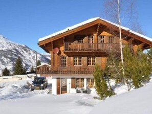 A two-story wooden chalet with a balcony, surrounded by snow and mountains. - Chalet Virginia A/B, (Grächen), by Interhome (Grächen)