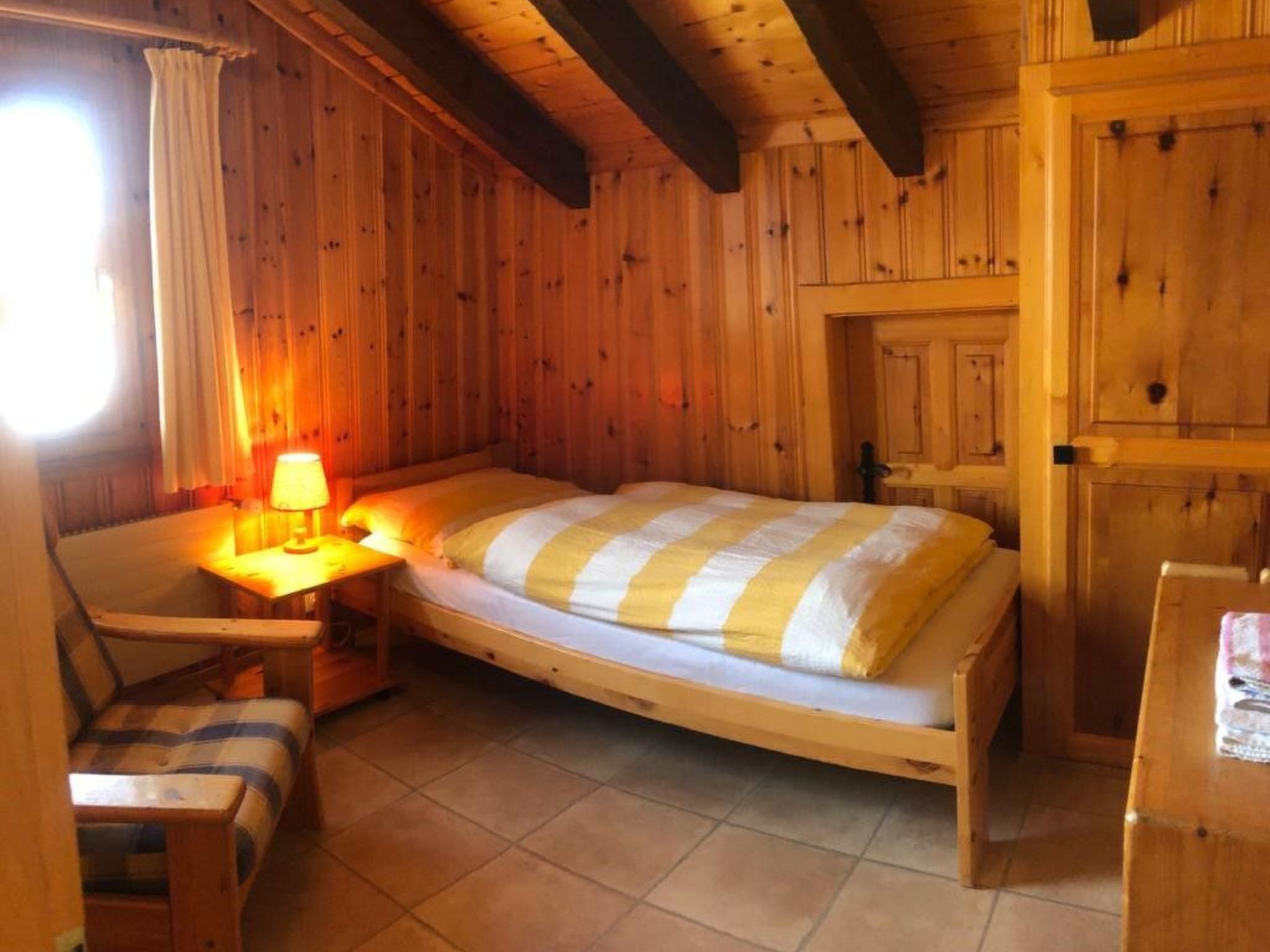 Una habitación de madera con una cama, una silla, una mesa y un ventanal con cortinas.