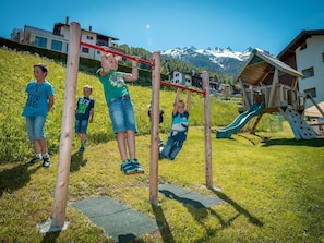 A playground with a wooden swing set and a slide, surrounded by grass and a mountain backdrop. - Ferienwohnungen Lunic, AZapartments.ch, B&B, (Grächen), 1057 by Interhome (Grächen)