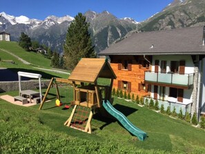 A playground with a slide and swings, a house with a balcony, and a mountain backdrop. - Ferienwohnungen Lunic, AZapartments.ch, B&B, (Grächen), 1057 by Interhome (Grächen)