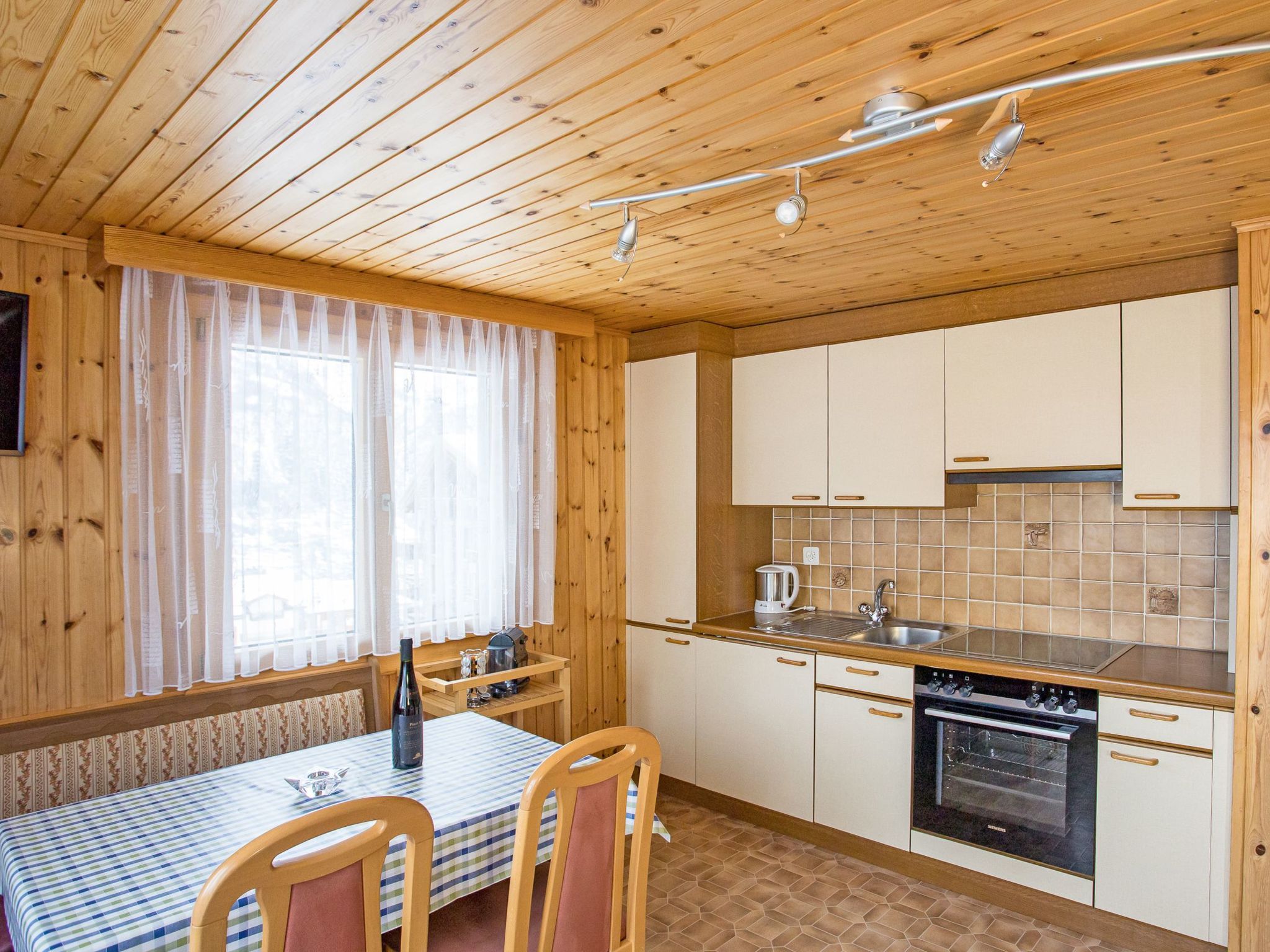 A wooden cabin interior with a dining area, kitchen, and a window with curtains.