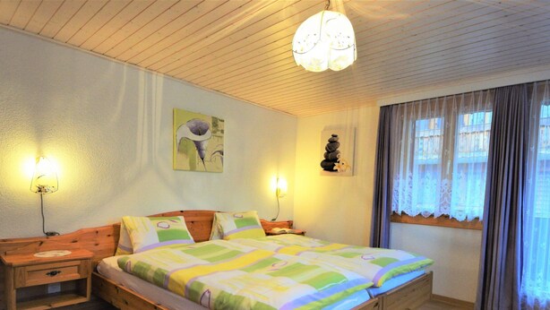 Een houten bed met een kleurrijke sprei, twee nachtkastjes met lampen, een wandklok en een raam met gordijnen.