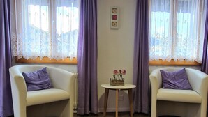 Twee witte fauteuils met paarse kussens, een klein rond houten tafeltje met een bloevas, en kantgordijnen voor de ramen.