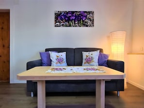 Quarto com sofá escuro, duas almofadas decorativas, mesa com toalha floral e um quadro de flores roxas fixado na parede.