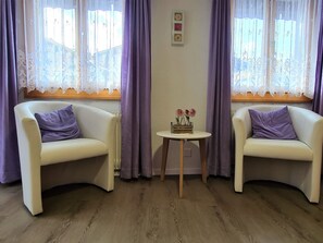 Ein Zimmer mit zwei weißen Sesseln, einem kleinen runden Tisch mit Blumenvase und einem Fenster mit violetten Vorhängen.