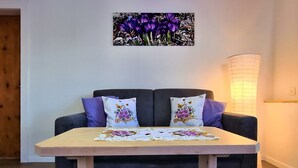 Een moderne woonkamer met een donkergrijze bank, twee paarse kussens, een tafelkleed met bloemenprint, en een aan de muur hangend schilderij van paarse bloemen.