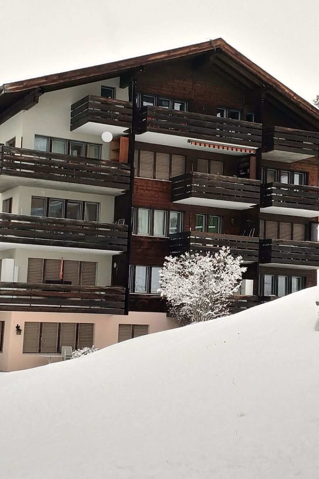 A multi-story building with balconies, large windows, and a snow-covered slope in the foreground.