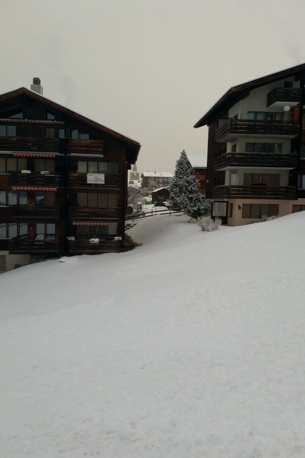 A snow-covered slope with wooden buildings, one with balconies and another with a flat roof.