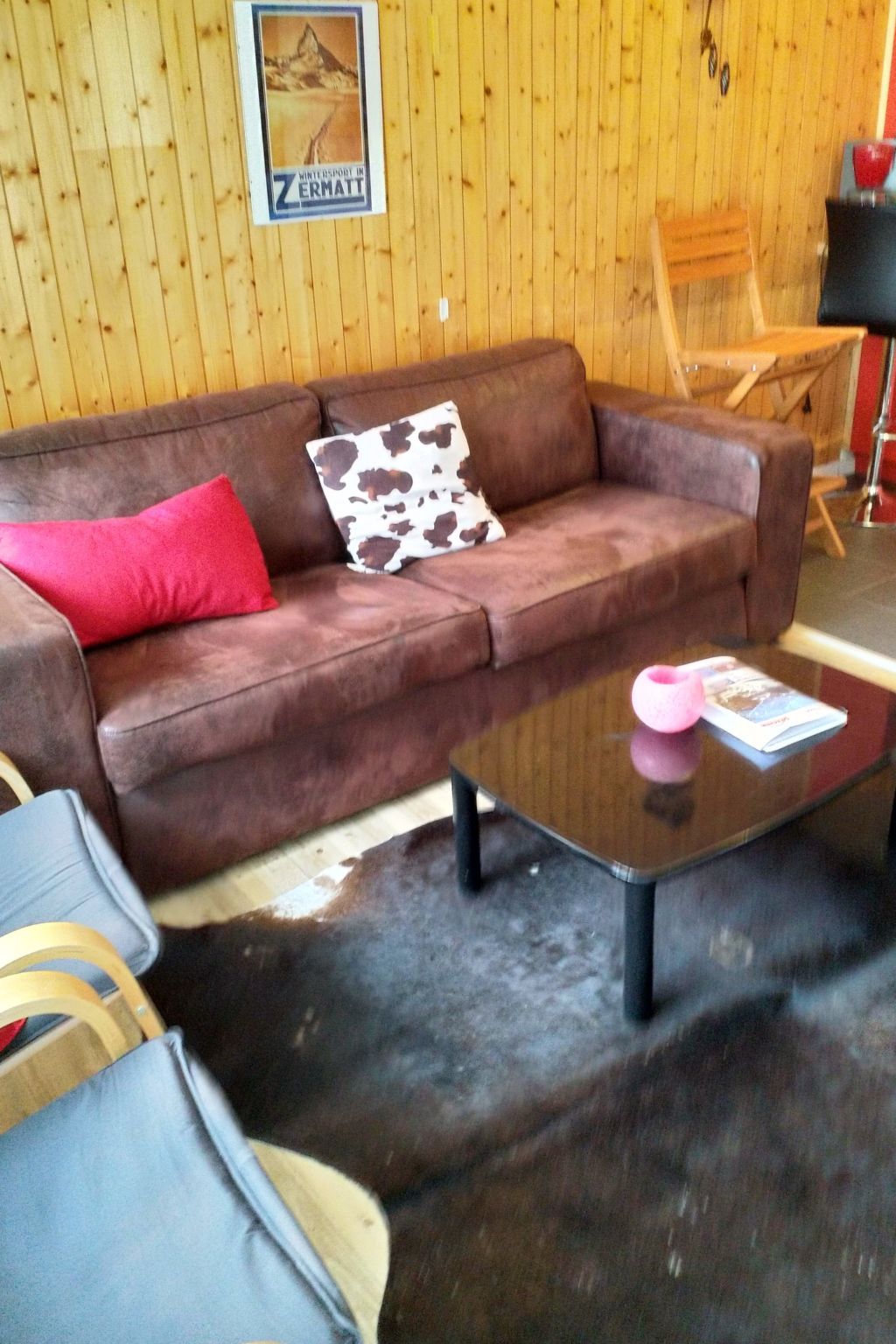 Un sofá de cuero marrón con un cojín rojo y uno estampado, una mesa de centro de vidrio con una revista, y una pared de madera con un cartel.