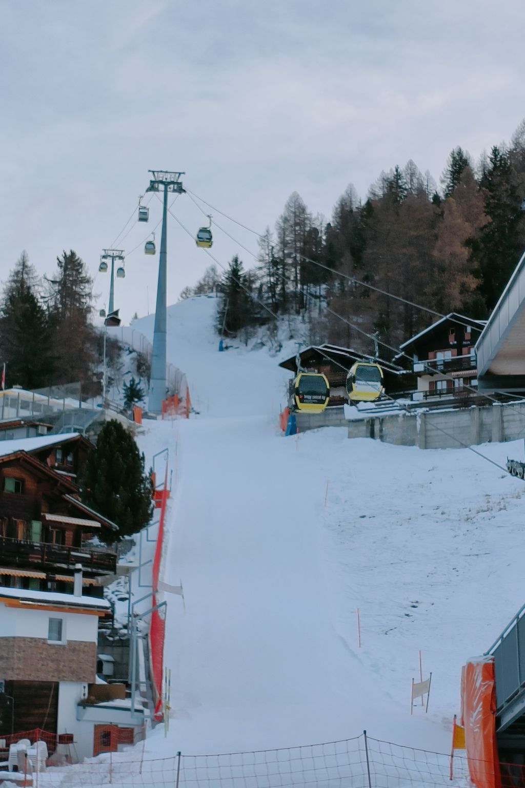 A ski lift with chairs, a snowy slope, and buildings in the background.