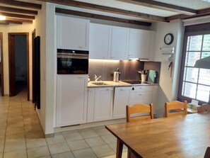 Eine kompakte Küche mit weißen Schränken, einer Spüle und einem Herd. Es gibt einen Esstisch mit Stühlen und ein Fenster mit Jalousien.