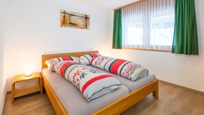 A single bed with a wooden frame and grey bedding, a bedside table with a lamp, a window with green curtains, and a painting on the wall. - AsterB, (Grächen), by Interhome (Grächen)
