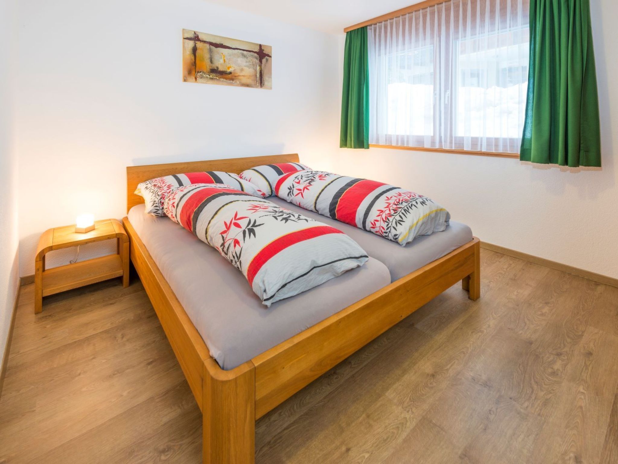 Ein Einzelbett mit Holzrahmen und grauem Lattenrost, ein Nachttisch mit Lampe sowie ein Fenster mit grünen Vorhängen.