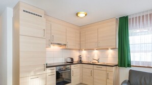 A compact kitchen with a built-in oven, a stovetop, and a sink. There is a green curtain on the window, and a leather chair is visible. - AsterB, (Grächen), by Interhome (Grächen)