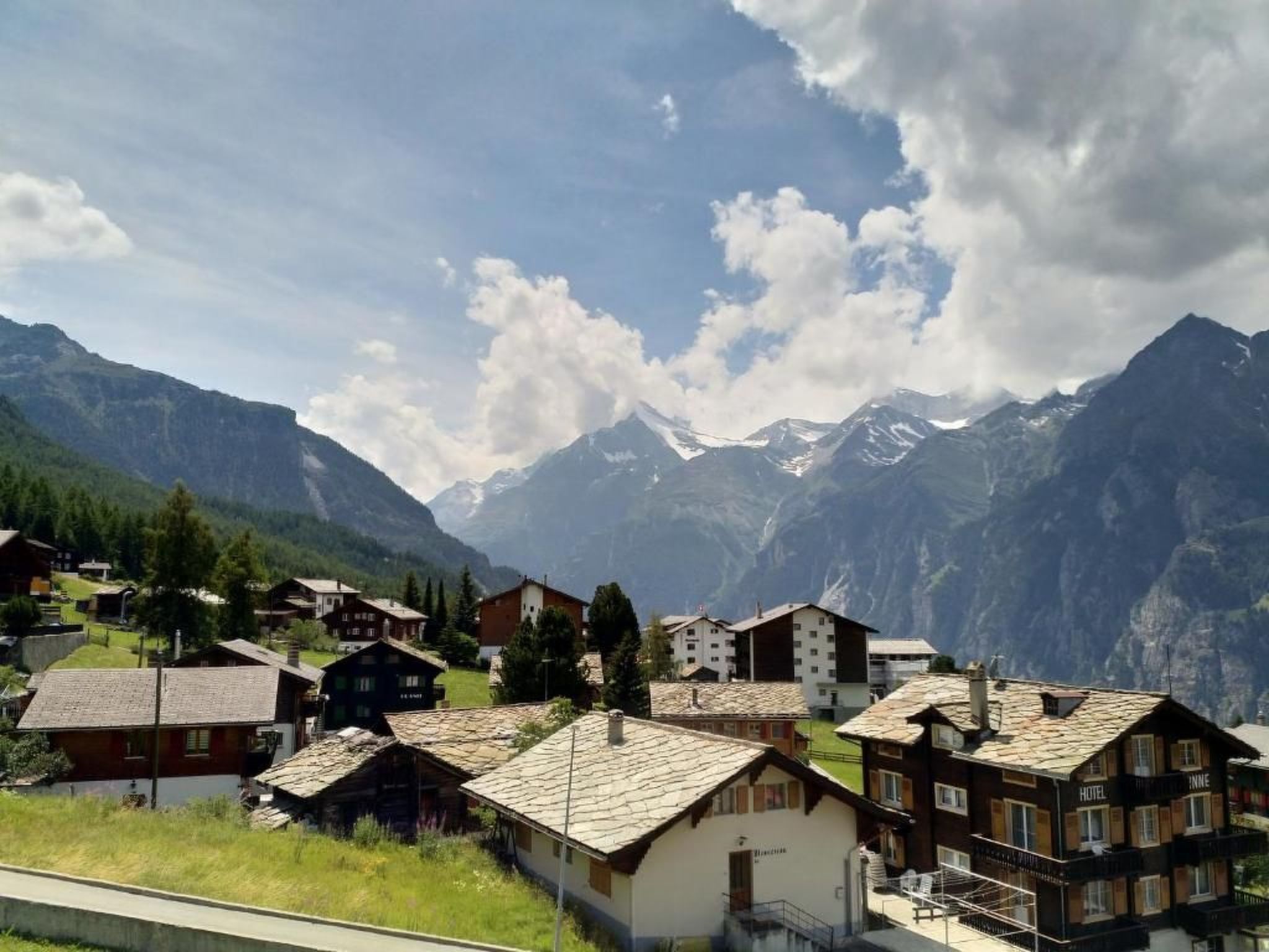 A mountain village with traditional chalet-style buildings, a clear sky, and snow-capped peaks in the background.