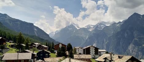 A mountain village with traditional chalet-style buildings, a clear sky, and snow-capped peaks in the background.