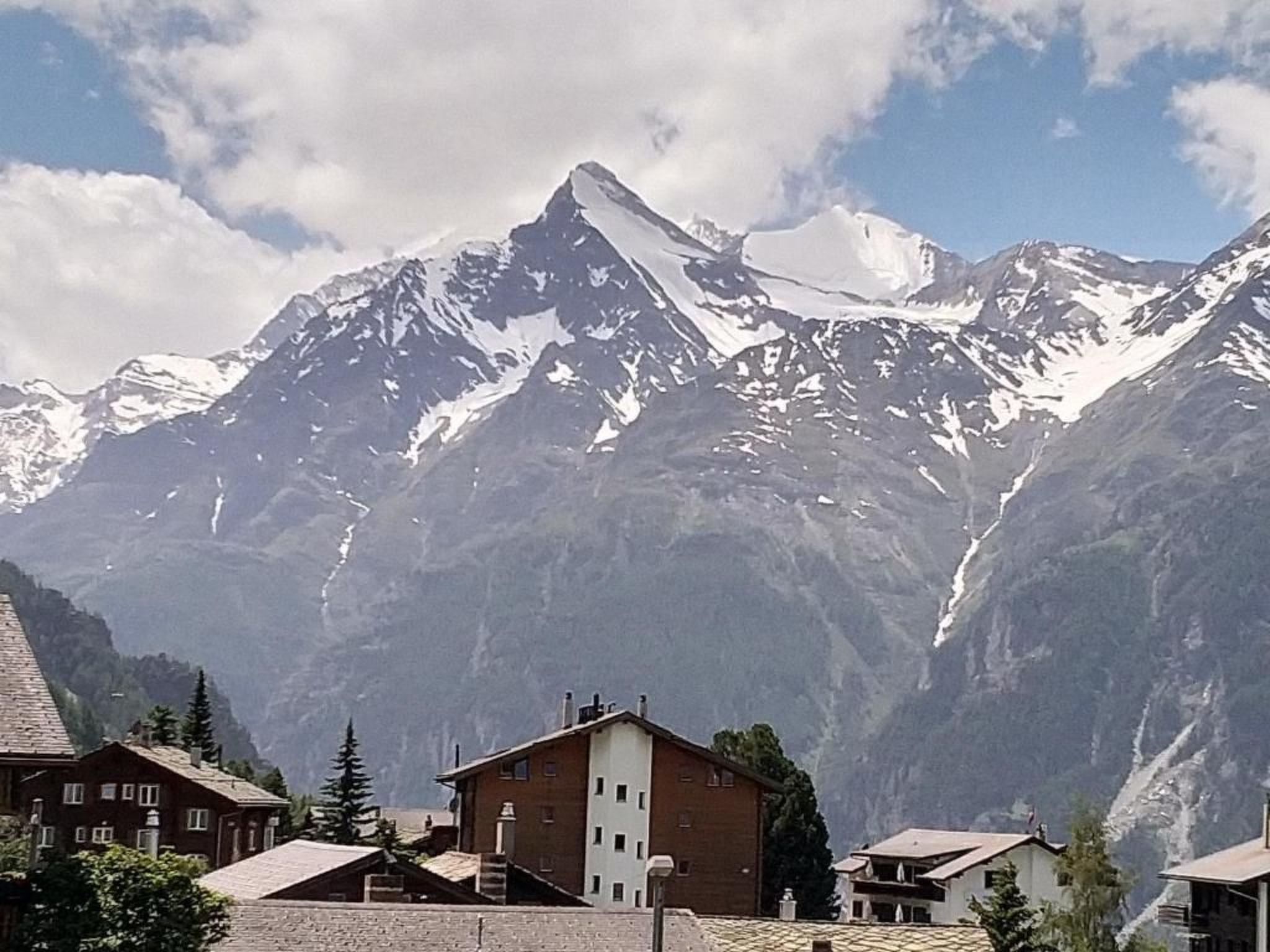 A mountain village with traditional chalet-style buildings, a prominent snow-capped peak in the background, and a clear sky.