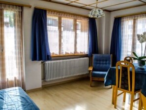 Ein Zimmer mit einem Bett, einem Tisch mit Stühlen, einem Heizkörper und einem Fenster mit Vorhängen.