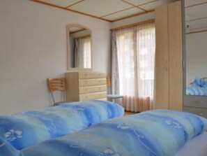 Ein Schlafzimmer mit zwei Betten, einem Schminktisch, einem Sessel, einem Spiegel und einem Fenster mit Vorhängen.