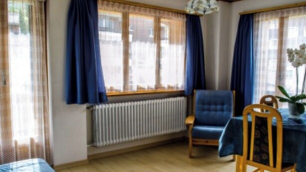 A room with a bed, a table with chairs, a radiator, and a window with curtains. - Alpfrieden, (Grächen), 1336 by Interhome (Grächen)