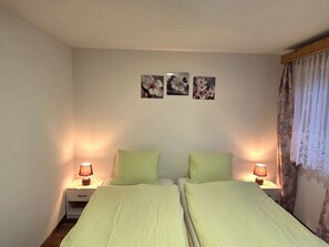 A double bed with two nightstands, each with a lamp, in a room with a plain wall and a window with curtains. - Ferienwohnungen Zur Heimat, (Grächen), by Interhome (Grächen)