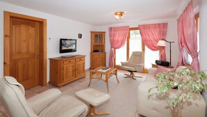 Ein Wohnzimmer mit Sofa, Sessel, Holzschrank, Fernseher und einer Zimmerpflanze.