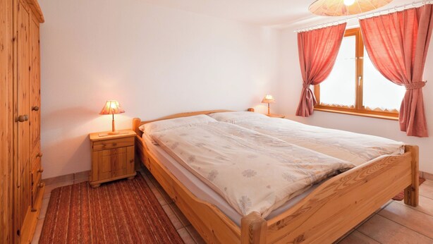 Ein hölzernes Bett mit einem gemusterten Überwurf, zwei Nachttischlampen, ein Holz-Nachttisch, ein gestreifter Teppich in Rot und ein Fenster mit rosa Vorhängen.