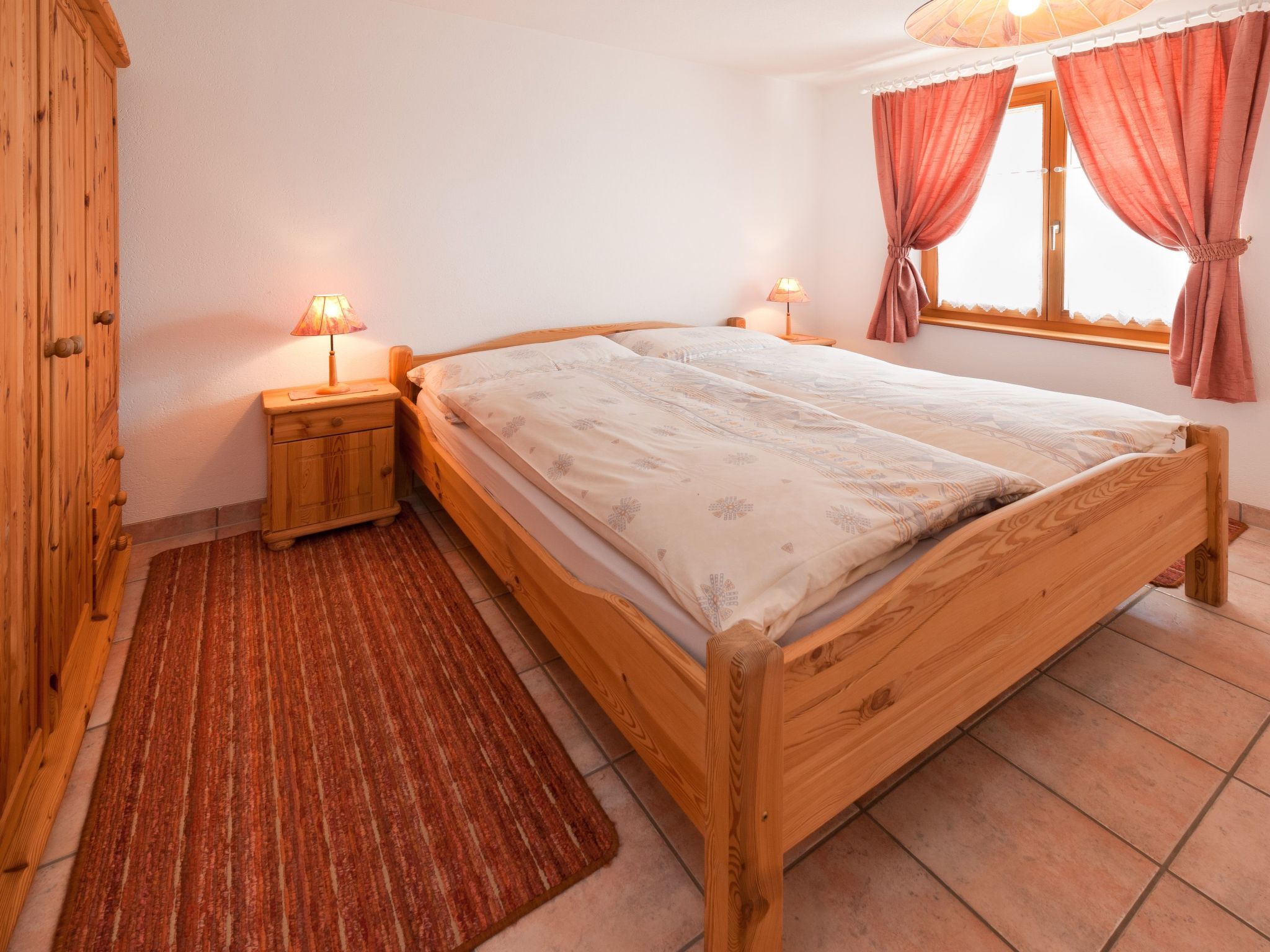 Un lit en bois avec une couvre-lit à motifs, deux lampes de chevet, une armoire en bois et un tapis rouge dans une pièce aux murs blancs et dotée d’une fenêtre avec des rideaux.