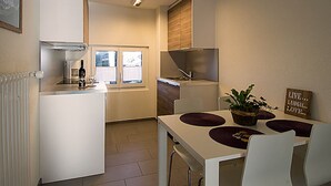 A compact kitchen with a refrigerator, stove, and window. There is a dining table with chairs and a potted plant. - Soldanella, (Grächen), by Interhome (Grächen)