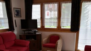 Ett vardagsrum med en röd soffa, en flätstol med en röd kudde, en tv på ett träställ och en mönstrad matta på golvet.