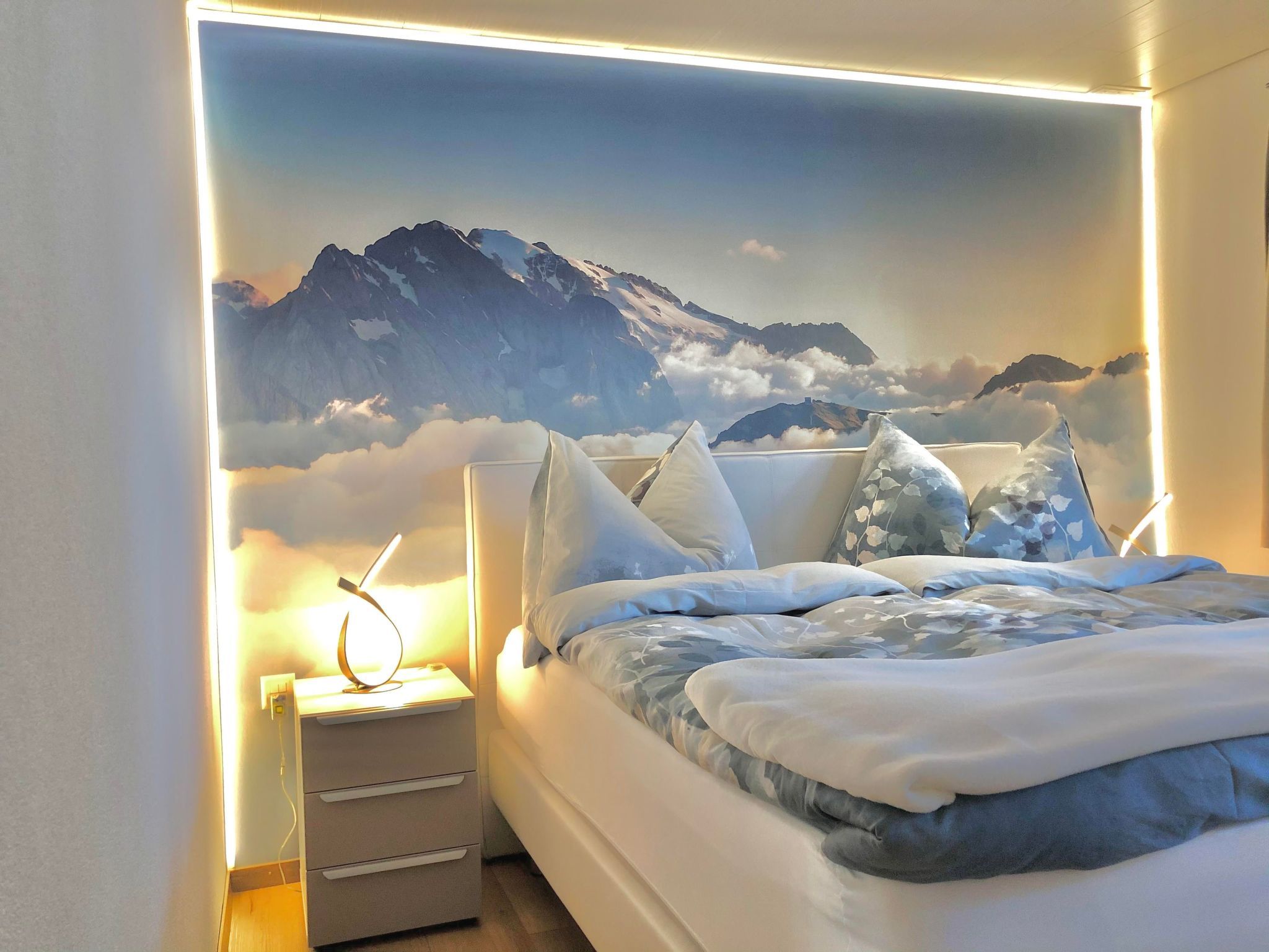 A bedroom with a mountain mural, a bed with white bedding, and bedside lamps.