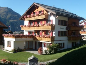 A multi-story hotel with balconies adorned with flowers, a sign reading 'Miranda,' and a mountainous backdrop. - Ferienwohnungen Miranda, (Grächen), by Interhome (Grächen)