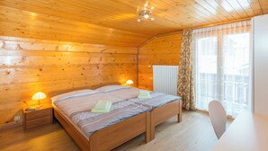 A wooden cabin room with a bed, bedside lamps, a table, and a window with curtains. - Ferienwohnungen Miranda, (Grächen), by Interhome (Grächen)