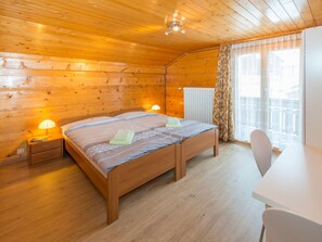 A wooden cabin bedroom with a bed, bedside lamps, a radiator, and a window with curtains. - Ferienwohnungen Miranda, (Grächen), by Interhome (Grächen)