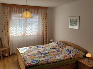 Une chambre à coucher avec un lit, une chaise, une table de chevet, une lampe et une fenêtre avec des rideaux.