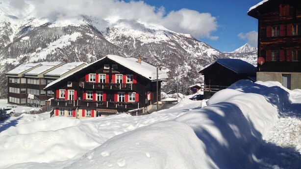 A snow-covered mountain village with traditional wooden buildings, including a chalet with balconies and a red facade. - Chalet Heimatglück, (Grächen), 1179 - 1181 by Interhome (Grächen)
