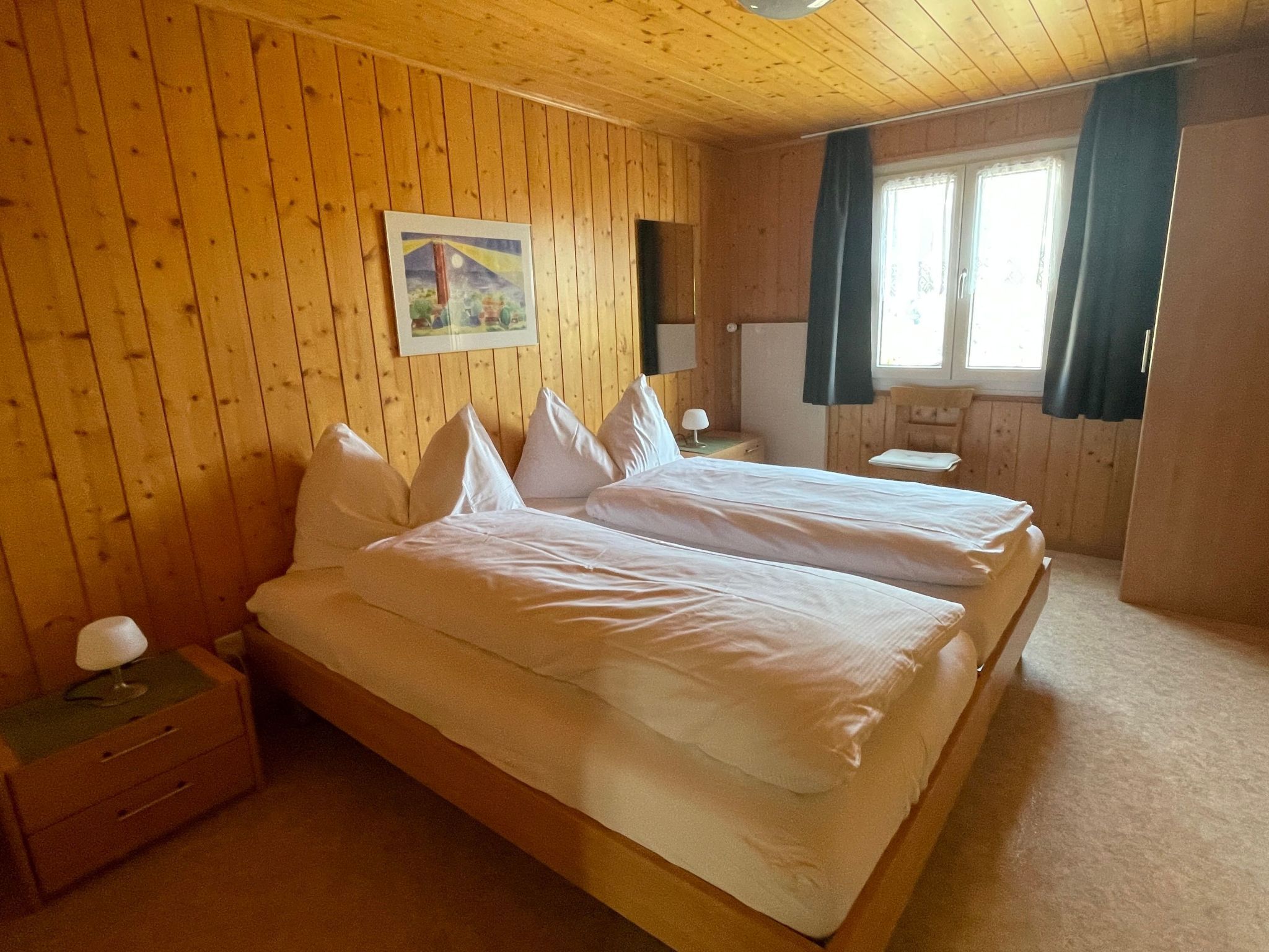 Een kamer met houten lambrisering, twee bedden, een nachtkastje, een lamp en een raam met gordijnen.