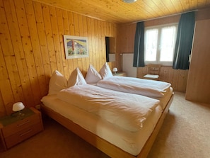 Ett rum med träpanel på väggarna, två sängar, ett nattduksbord, en lampa och ett fönster med gardiner.