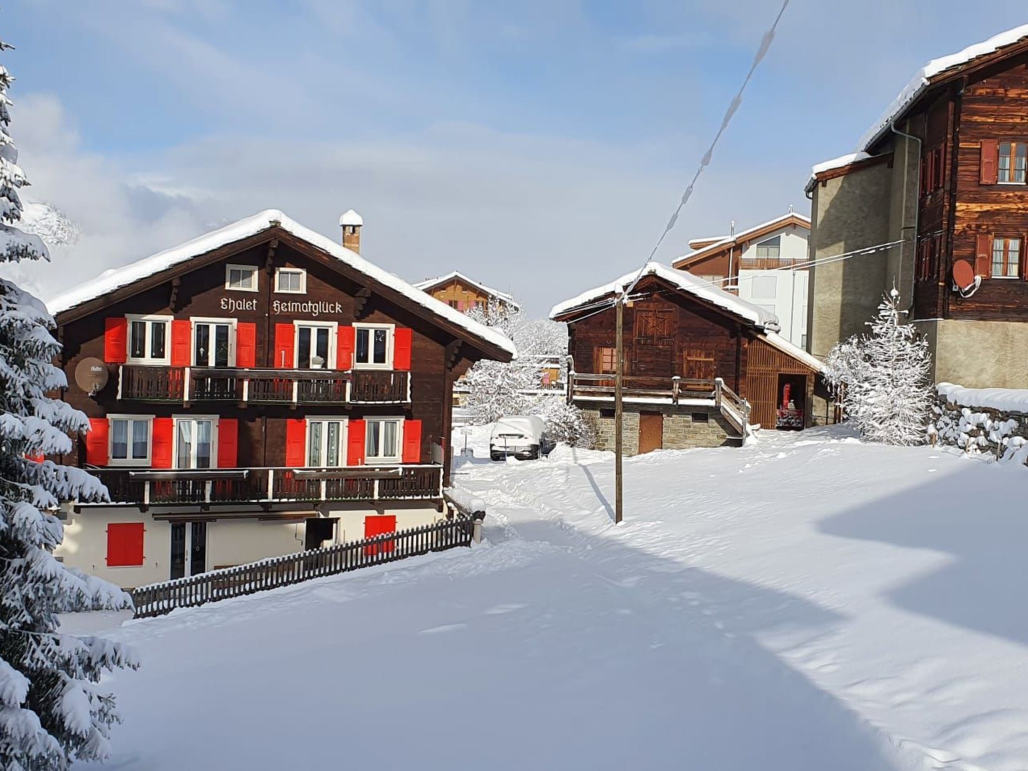 Ein Chalet mit Balkon und roten Fensterläden, umgeben von schneebedeckten Bäumen und blauem Himmel.