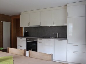A modern kitchen with white cabinetry, a built-in oven, and a dining area with a table and chairs. - Chalet Heimatglück, (Grächen), 1179 - 1181 by Interhome (Grächen)