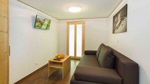 A small living room with a brown sofa, a wooden side table with a fruit bowl, a television, and a door leading to another room.