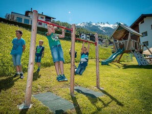 A playground with a wooden swing set and a slide, surrounded by grass and a mountain backdrop. - Ferienwohnungen Lunic, AZapartments.ch, B&B, (Grächen), 1057 by Interhome (Grächen)