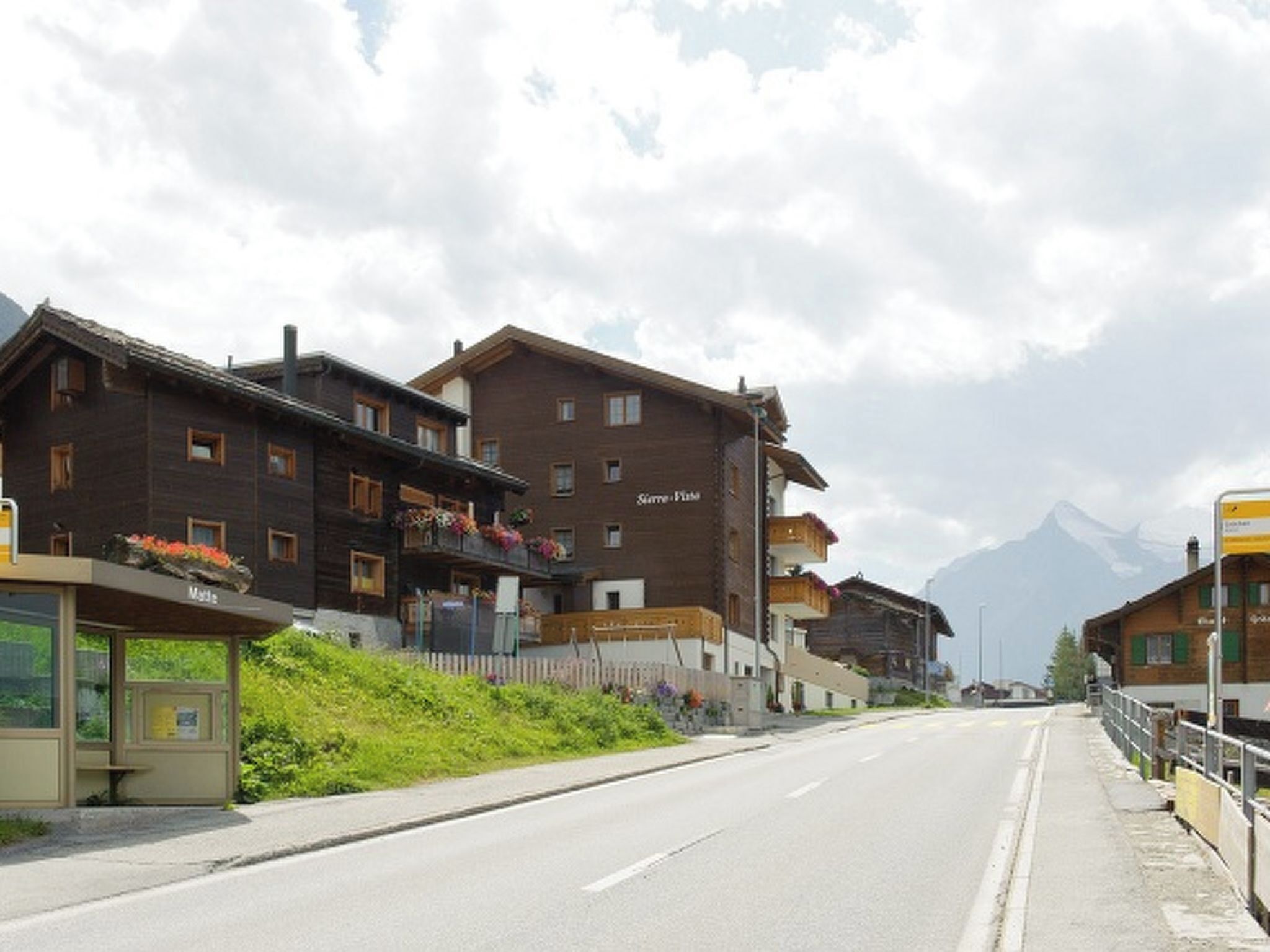 Straßenansicht mit traditionellen Holzhäusern, blauem Himmel und einem Berg im Hintergrund.